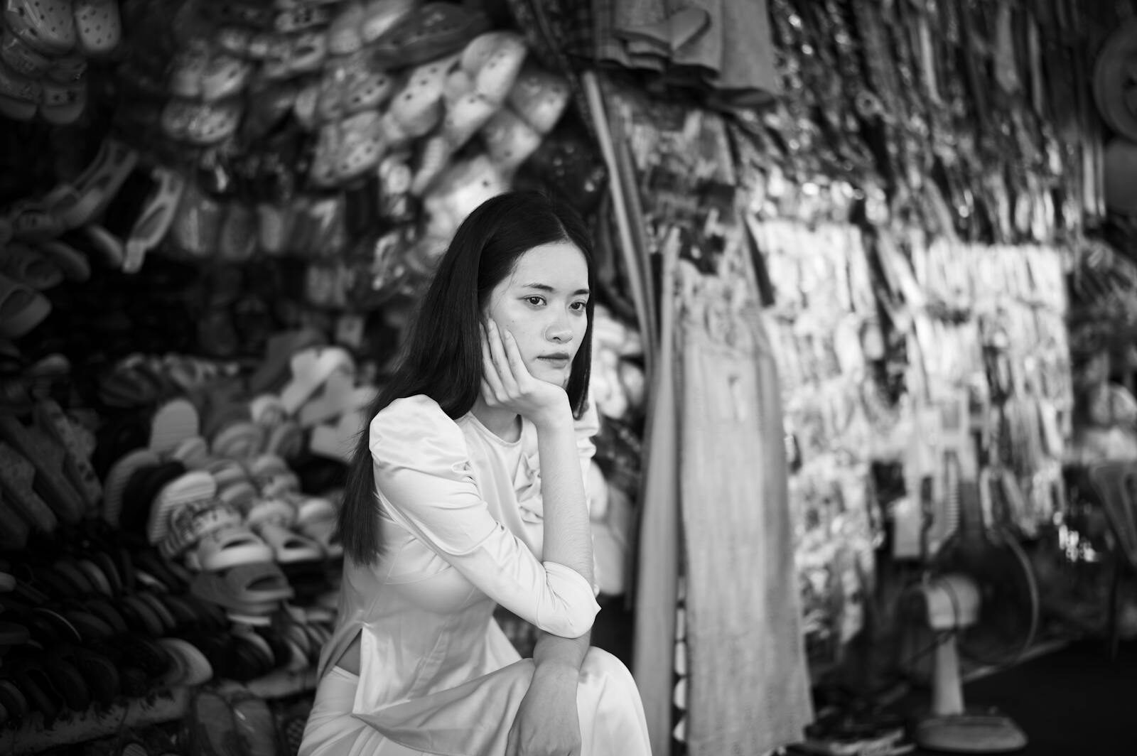 Black and white portrait of a woman sitting thoughtfully in an Asian market with various goods.