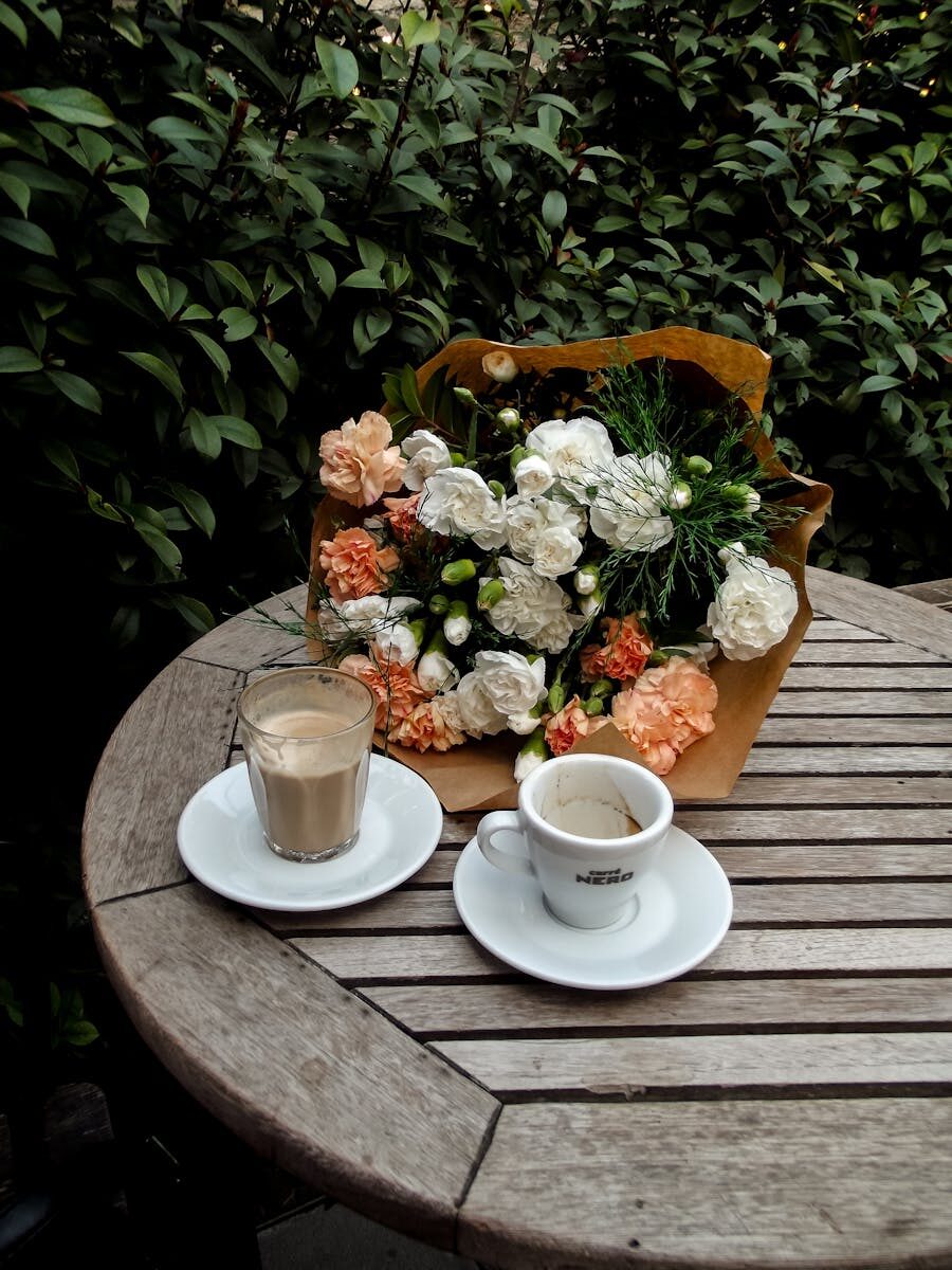 Wooden table with coffee cups and a bouquet, set in a lush garden setting.